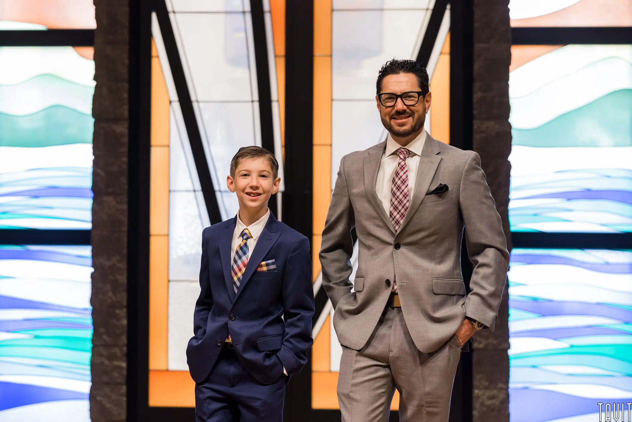 A boy and a man wearing suits and ties stand smiling in front of colorful stained glass windows at a corporate event.