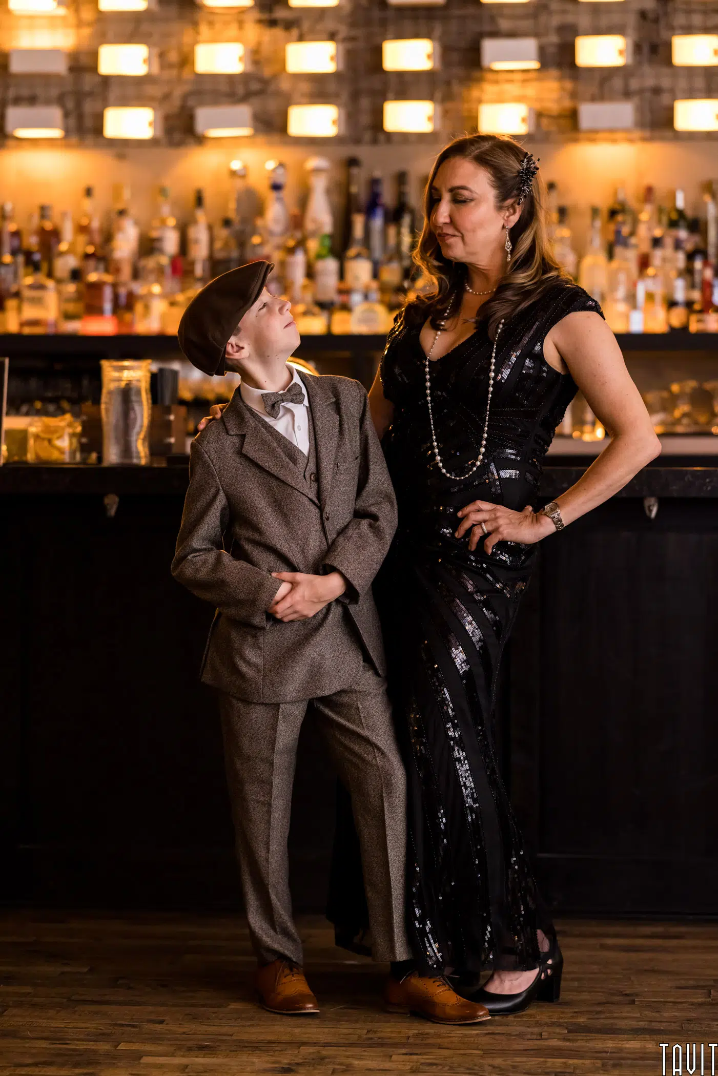 A woman in a black sequined dress and a boy in a vintage grey suit and cap stand together in front of a bar at a corporate event, looking at each other and smiling, with warm lighting in the background.