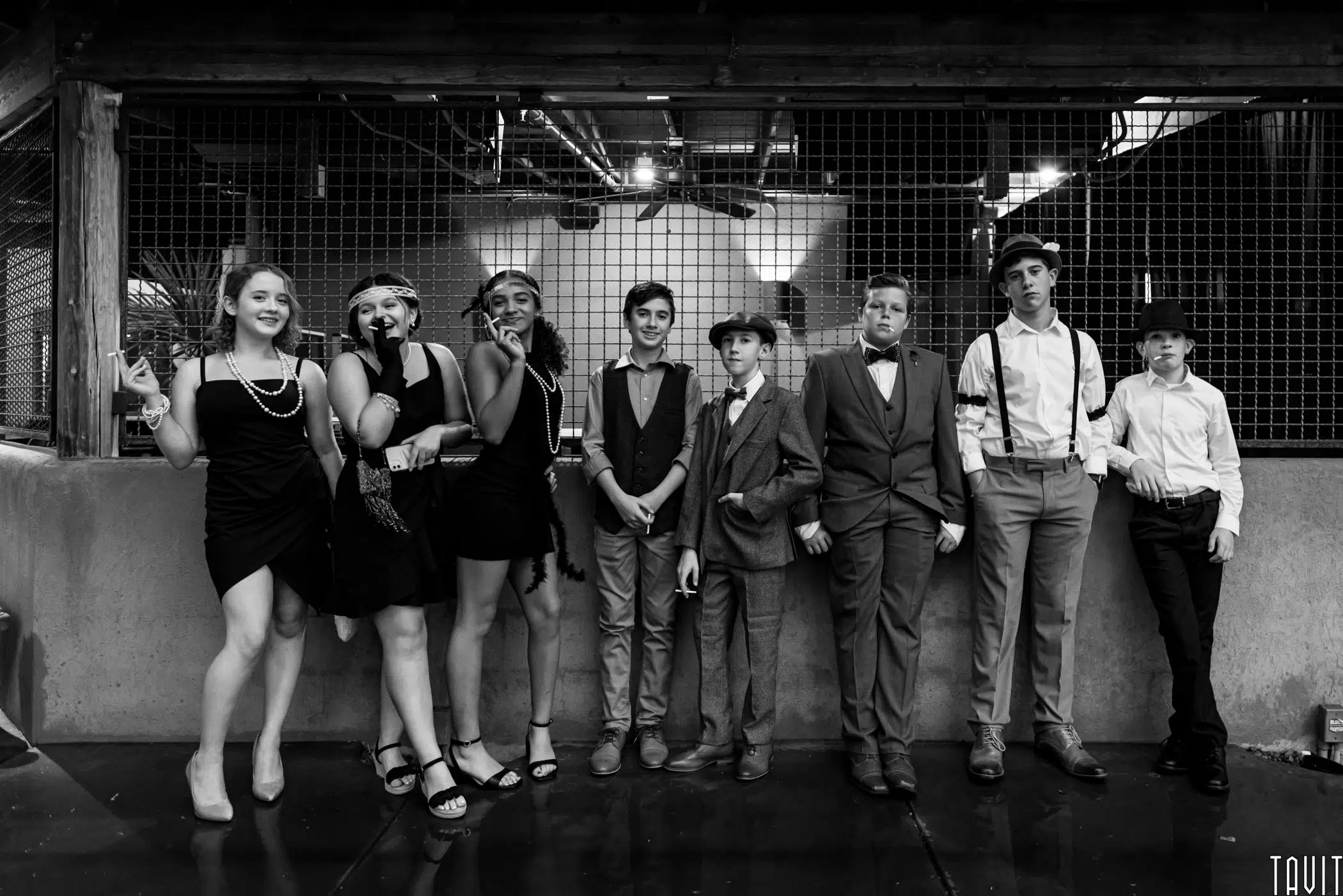 A group of eight teens dressed in 1920s-style outfits pose together at a corporate event, some smiling, some serious, in front of a wire fence in a dimly lit setting. The photo is in black and white.
