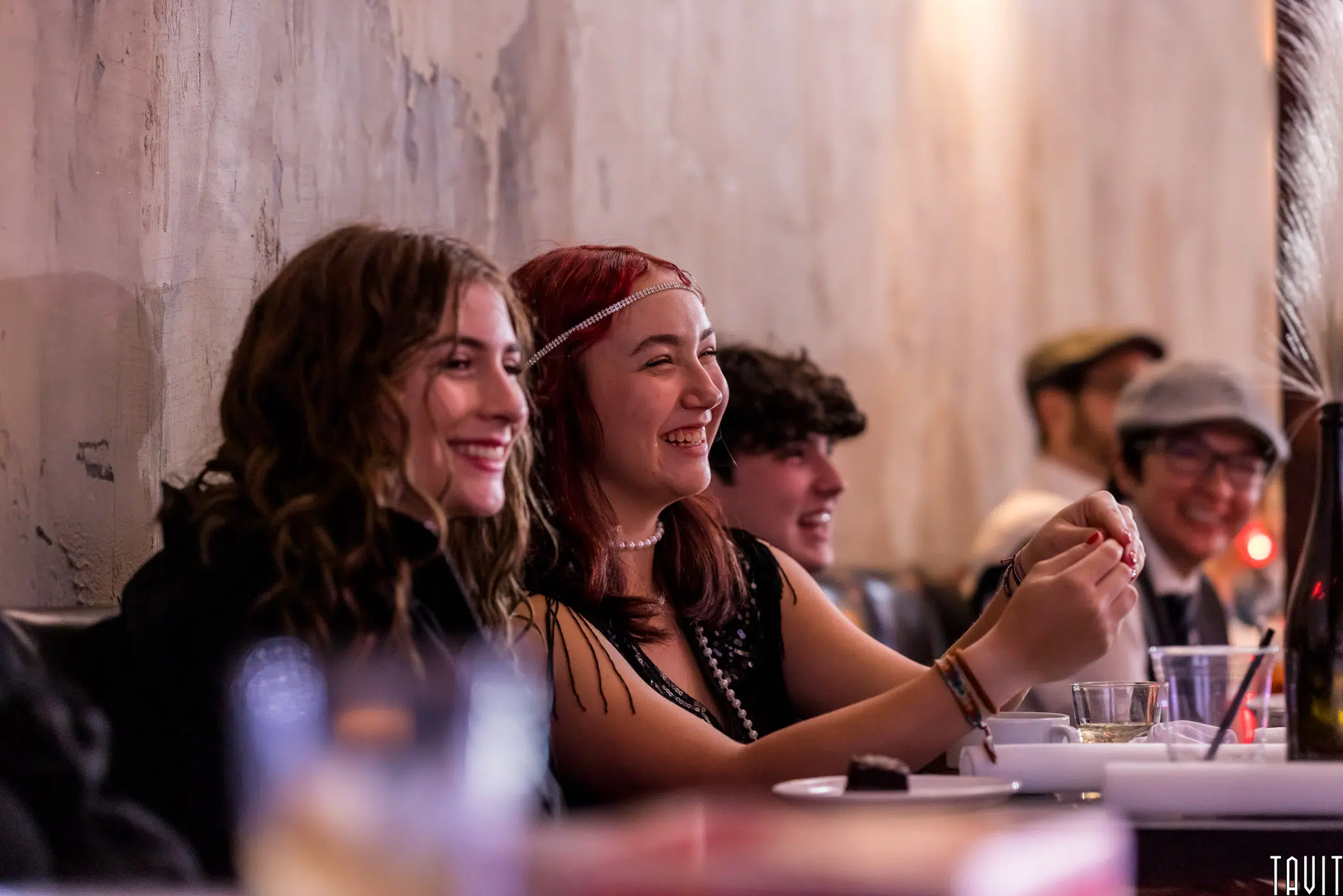 Three young people sit together at a table, smiling and laughing. The group appears to be enjoying themselves in a warmly lit, casual indoor setting at a corporate event with food and drinks in front of them.
