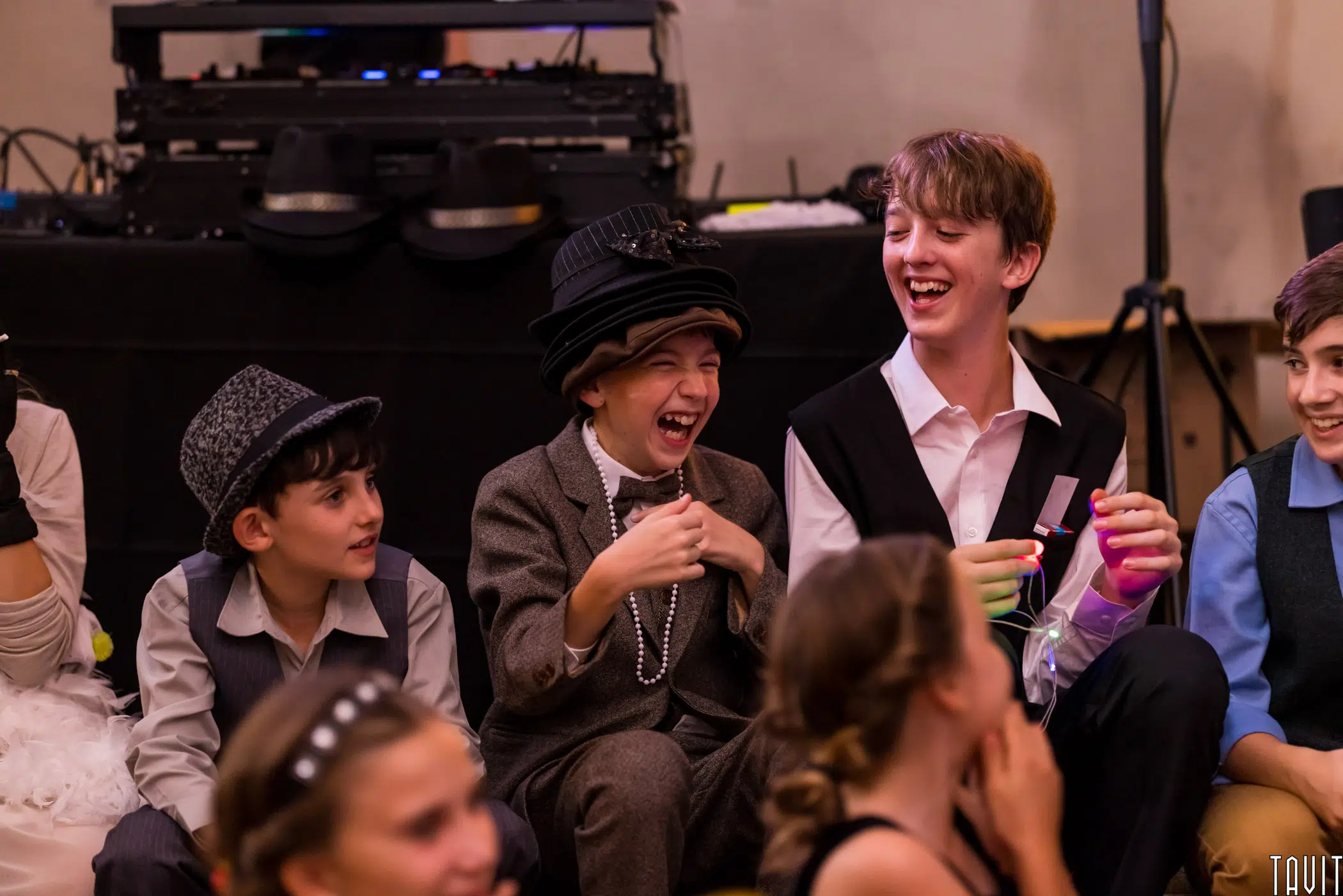A group of kids dressed in vintage-style costumes sit on the floor, laughing joyfully. In the background, DJ equipment hints at a lively expo or festive corporate event atmosphere.