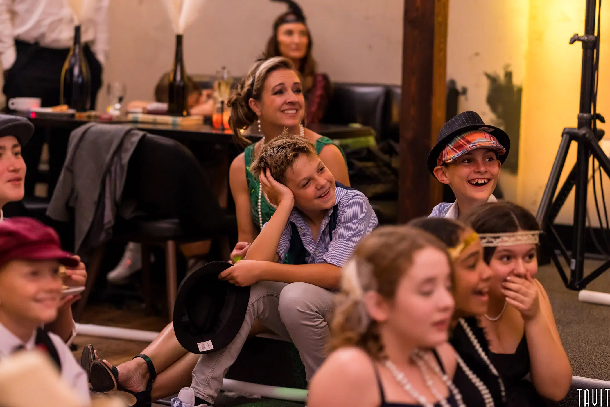 A group of children dressed in vintage or 1920s-style clothing sit on the floor, laughing and smiling during a seminar. Behind them, an adult woman also smiles. The background includes a table with drinks and decorations.