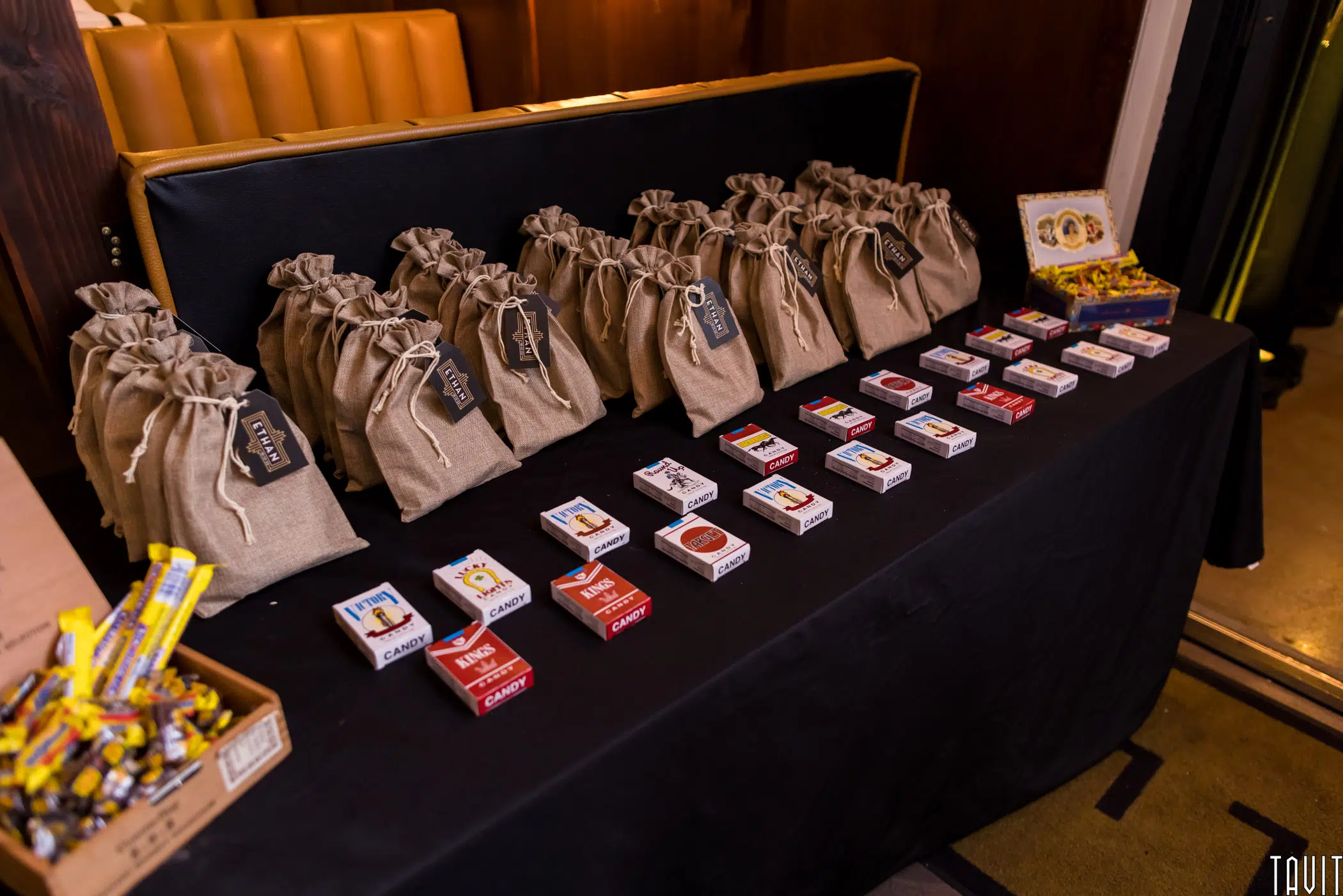 A table displays rows of matchboxes with various designs, burlap gift bags labeled with tags, and a pile of yellow-wrapped candies, set in a warmly lit indoor setting at a corporate event.
