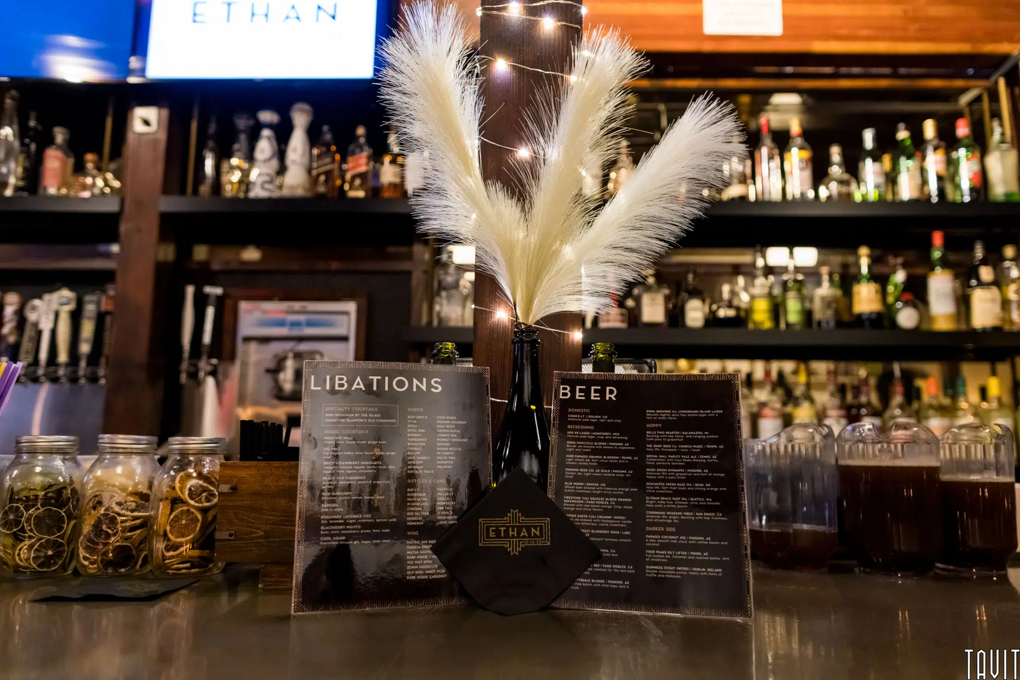 At a corporate event, a bar counter displays cocktail ingredients in jars, two drink menus labeled “Libations” and “Beer,” a black napkin with “Ethan” in gold, and a vase with white feathery decor, with liquor bottles in the background.