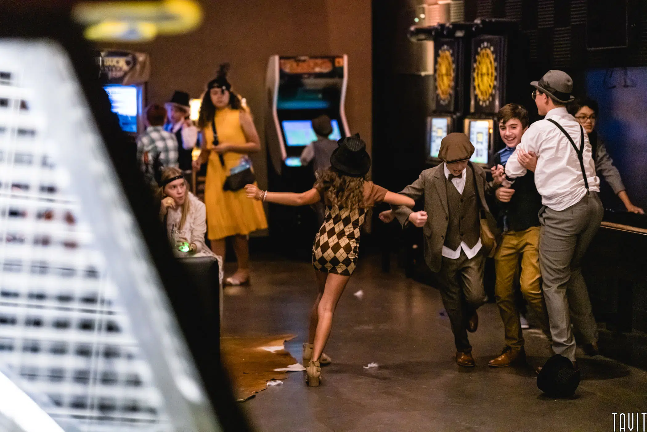 A group of people in vintage-style outfits dance and play at an arcade-themed corporate event. Some stand near arcade machines, while others hold hands and laugh in the center of the room, creating a lively and festive atmosphere.