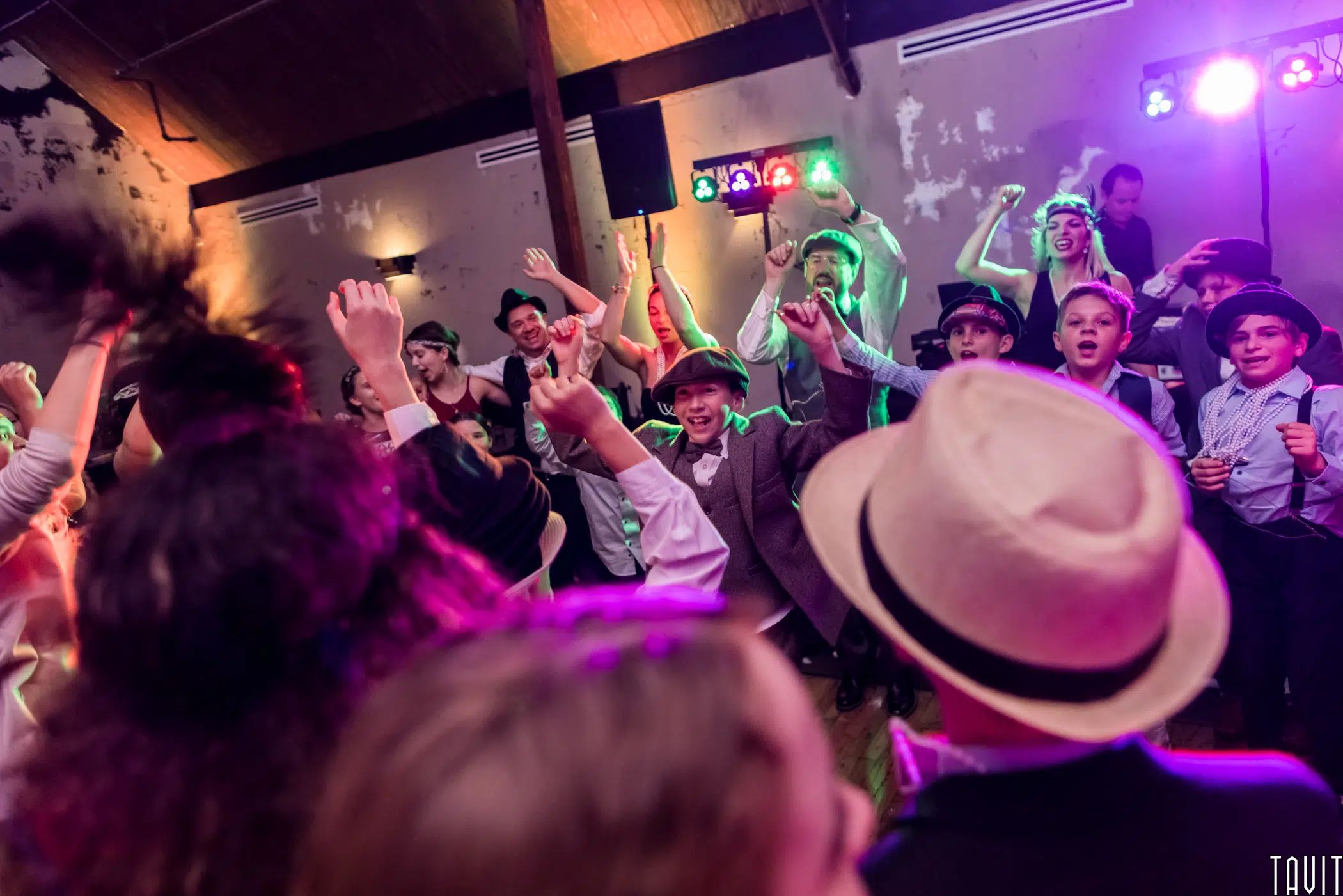A lively group of people, many wearing hats and costumes, dance energetically under colorful lights at an indoor corporate event. The crowd has raised hands and joyful expressions in a rustic venue with exposed wooden beams.
