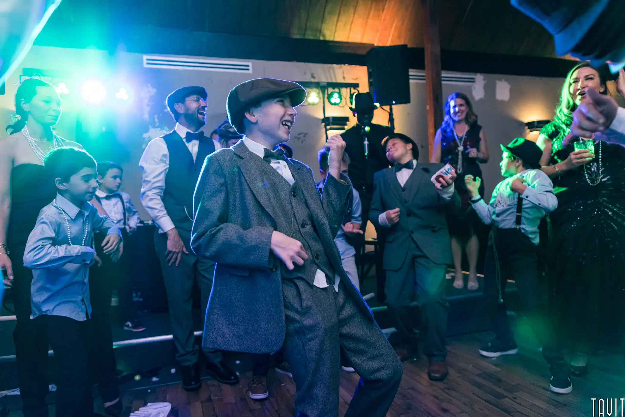 A young boy in a vintage suit and cap dances enthusiastically at a lively indoor corporate event, surrounded by children and adults in festive attire under colorful lights.