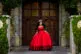 young lady at her quinceañera, in a red and black dress, standing in front of wooden doors.