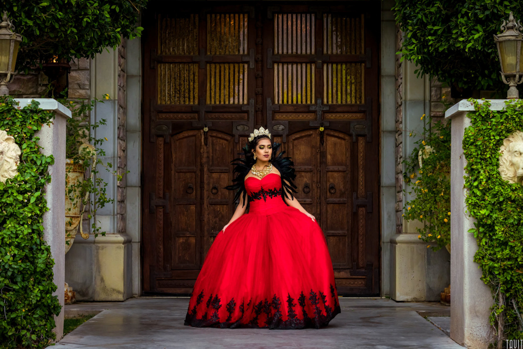 young lady at her quinceañera, in a red and black dress, standing in front of wooden doors.
