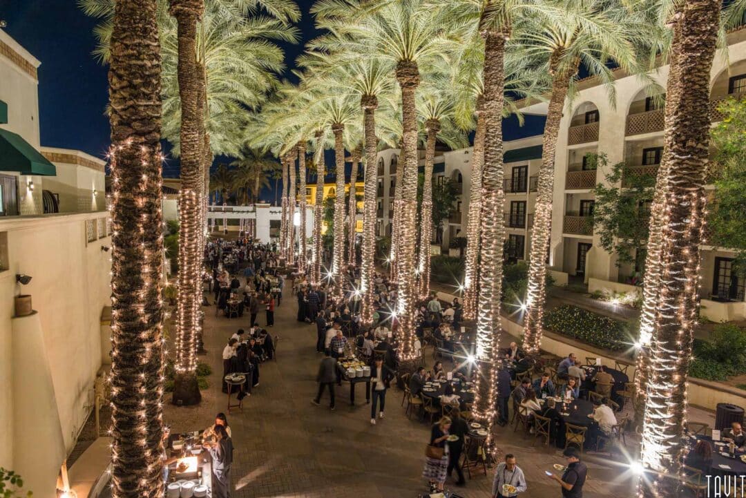 A nighttime event with people mingling under rows of tall palm trees wrapped in string lights. Tables with food and drinks are set up along the path, and a multi-story building with arched windows is visible in the background.