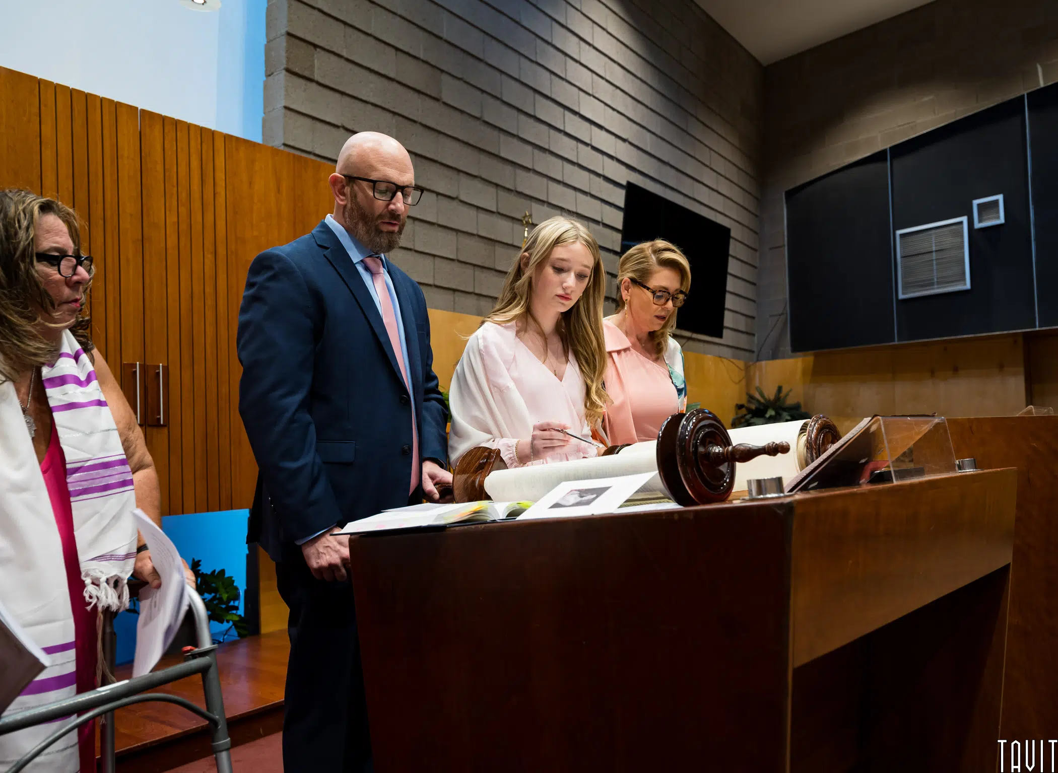 A person stands at a lectern, reading from a book, accompanied by three others. They are in a room with wooden paneling and a brick wall. Two men and two women are present, dressed formally, focusing on the event.