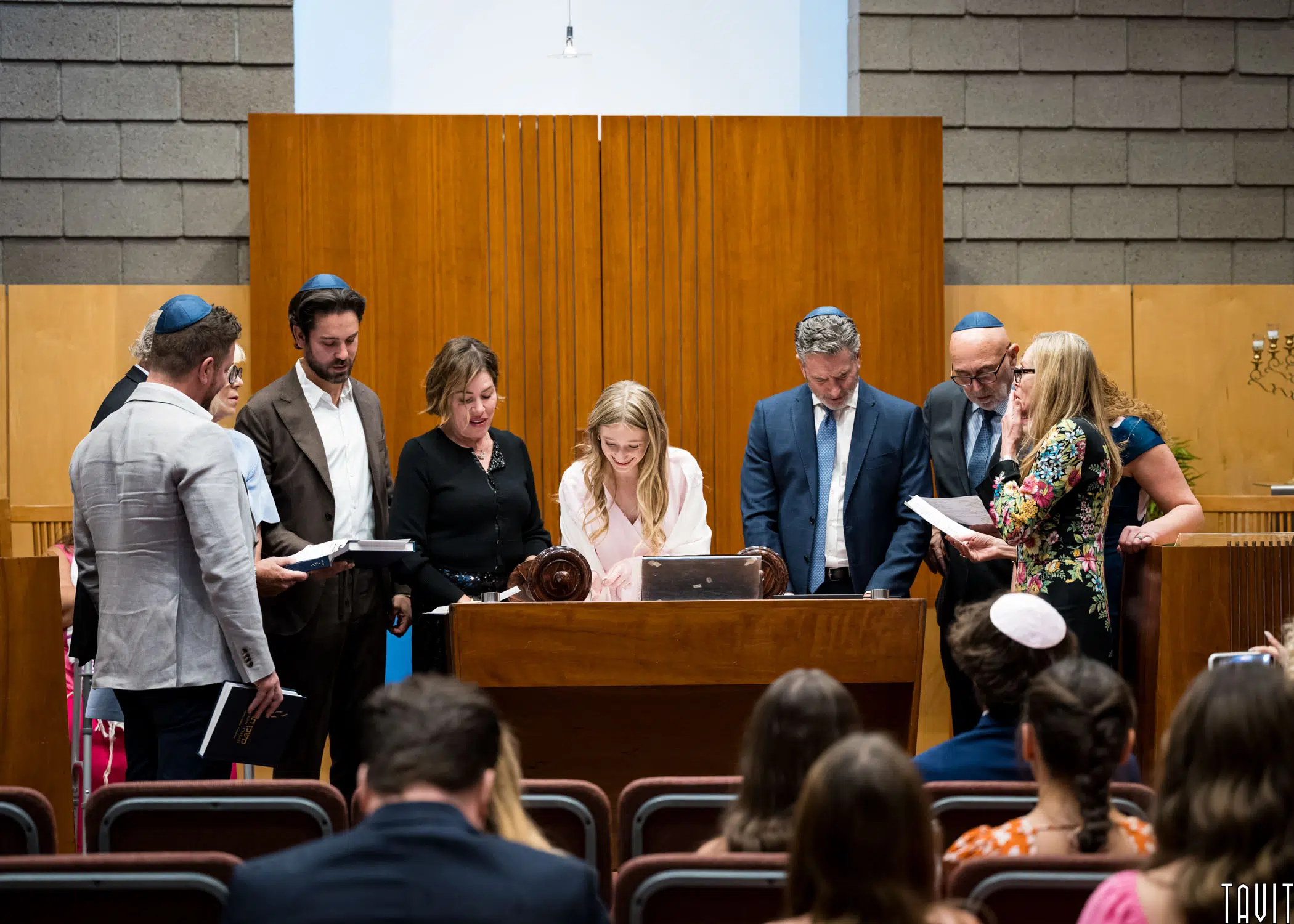 A group of people gathered around a girl reading from a Torah scroll in a synagogue. The setting features wooden paneling and guests seated in the foreground. Some individuals wear kippahs. Everyone is focused on the central activity.