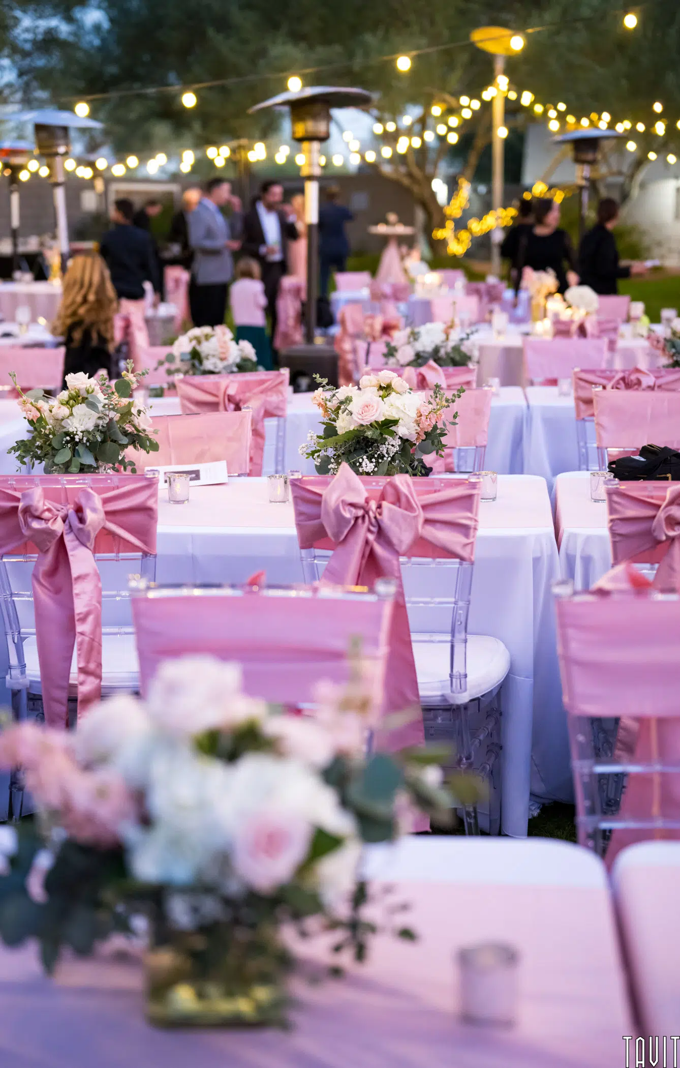 Outdoor wedding reception with tables and chairs adorned in pink covers and bows. Each table features floral centerpieces with white and pink roses. String lights hang above, creating a warm and festive atmosphere.