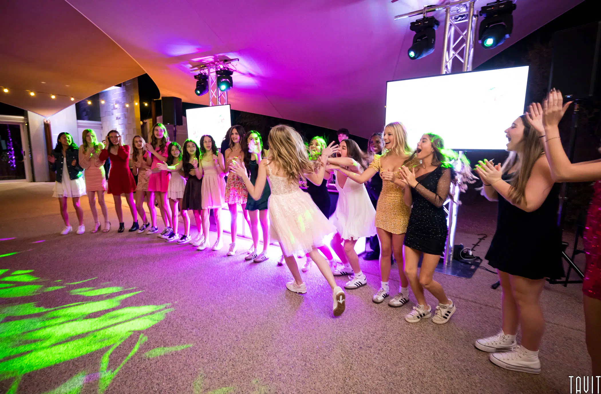 A group of young women in colorful dresses and sneakers stand in a semicircle, clapping and cheering under vibrant lights at an outdoor event. One girl in a white dress joyfully runs towards the group.