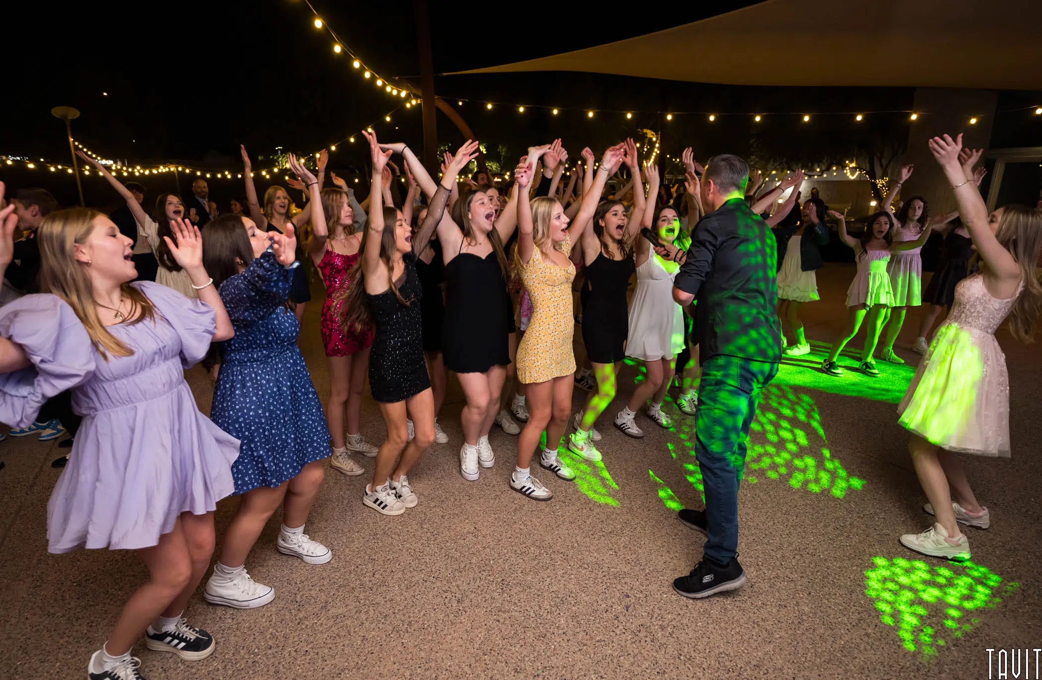 A group of people dance energetically in an outdoor setting with string lights overhead. Many are wearing casual clothes and sneakers. There is a mix of colorful outfits, and green lighting creates dynamic patterns on the ground.