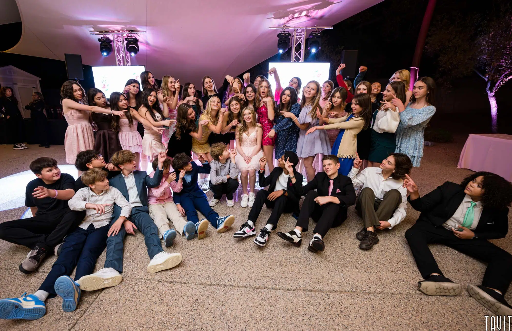 A large group of young people at a lively event, some seated on the ground and others standing. They're dressed in formal and semi-formal attire, with lots of laughter and playful gestures. The setting is decorated with colorful lights.