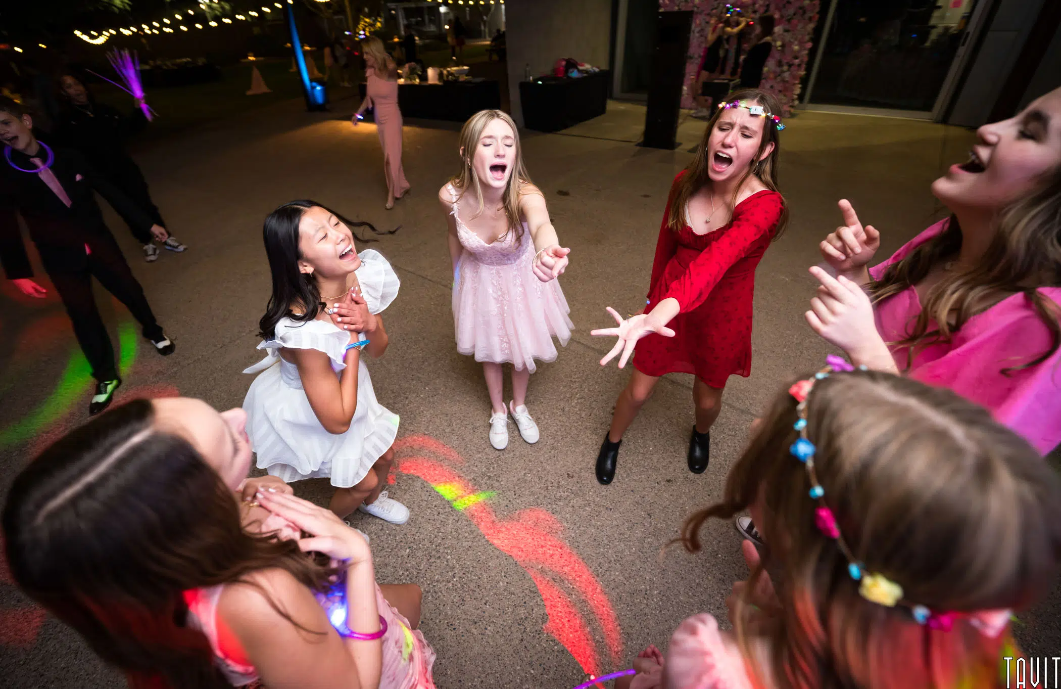 A group of young people dressed in bright party clothes dance energetically in a festively lit space. They appear joyful and animated, with some reaching out or singing. Strings of lights and colorful reflections add to the lively atmosphere.