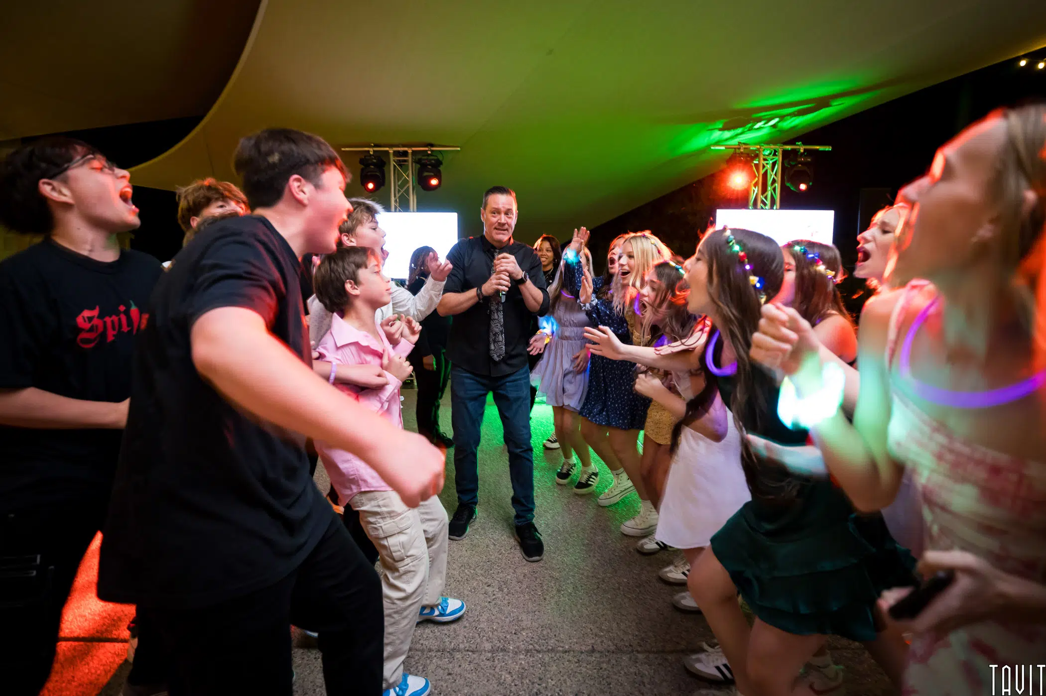 A lively scene of a group of people dancing and enjoying themselves at a party. A person with a microphone stands at the center, engaging the crowd under colorful lights and a decorated ceiling.