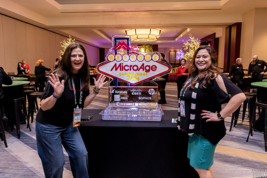 Two women smiling and posing next to a large ice sculpture shaped like a casino sign that reads "Welcome MicroAge Casino Night 2024." The venue is decorated with illuminated trees and gaming tables in the background.