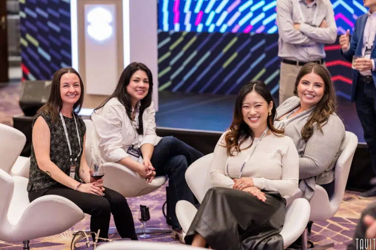 Four women seated in white chairs at a conference, smiling and looking towards the camera. The background features a colorful, abstract display. The setting appears to be indoors with modern decor.