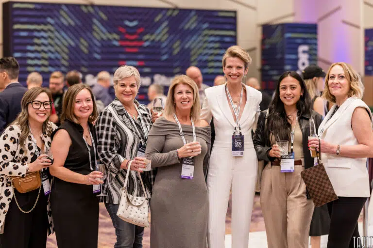 A group of seven women stands together at an event, smiling for the camera. They are dressed in a mix of casual and business attire, wearing name tags. The background features other attendees and colorful decor.
