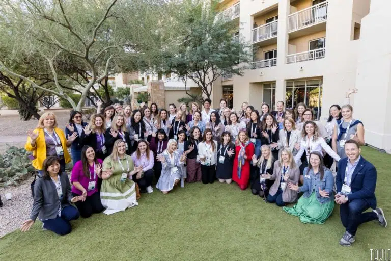 A large group of people, mostly women, pose together on a grassy area in front of a building and trees. They are smiling and waving at the camera, wearing diverse outfits and name badges, suggesting a conference or professional event.