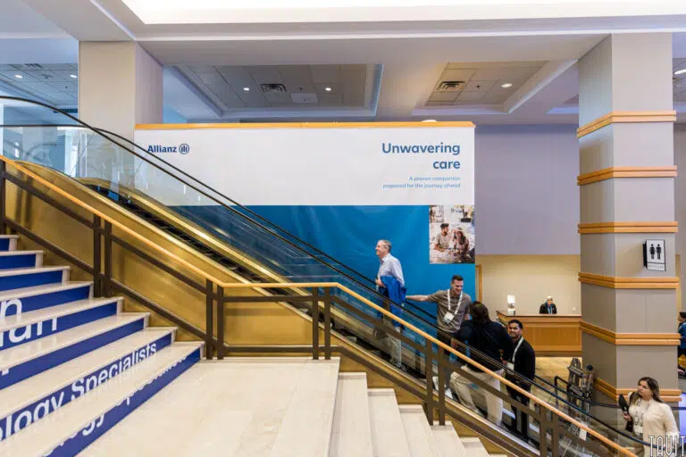 A staircase in a conference venue with a banner displaying the Allianz logo and the text "Unwavering care." People are ascending and descending the stairs. The surroundings include beige walls and a wooden railing.