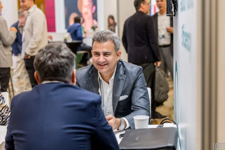 Two men in a professional setting are engaged in conversation at a conference or business meeting. One man faces the camera, smiling, with a coffee cup and laptop on the table. Other people and booths are seen in the blurred background.