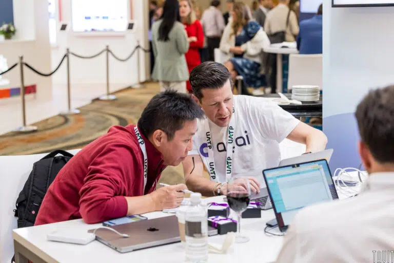 Two people sitting at a table with laptops, focusing on a project. One wears a red sweater and the other a white shirt with "ai" visible. A glass of red wine and water bottle are on the table. Others are in the background at a busy event.