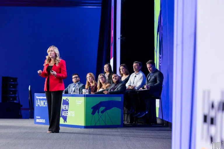 A woman in a red blazer is speaking on a stage with a panel of eight people seated at a table behind her. The background displays vibrant graphics and the words "Launch" and "New Agents." The setting appears to be a conference or presentation.