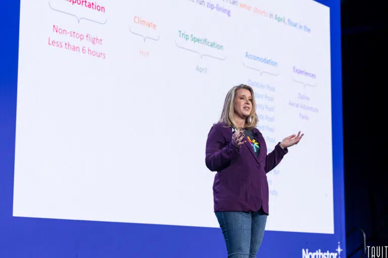 A person stands on stage giving a presentation. They wear a dark shirt and jeans, gesturing with one hand. A large screen behind them displays a slide with colorful text about transportation, climate, trip specifications, and experiences.