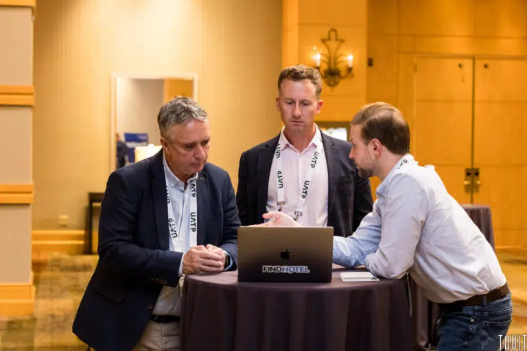 Three men in business attire stand around a round table, focused on a laptop screen with "FINDHOTEL" on it. They are in an indoor setting with warm lighting and visible conference badges.