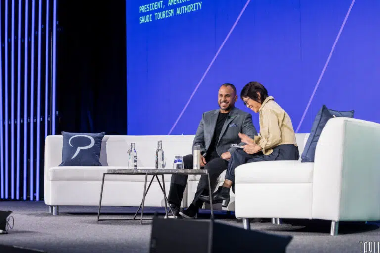 Two people are seated on a stage with white couches and a small table, engaged in conversation. The background features a large screen displaying text. Water bottles and a microphone are on the table.