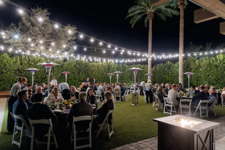 An outdoor evening event with people seated at round tables under string lights. The scene features palm trees, patio heaters, and greenery, creating a warm, inviting atmosphere.