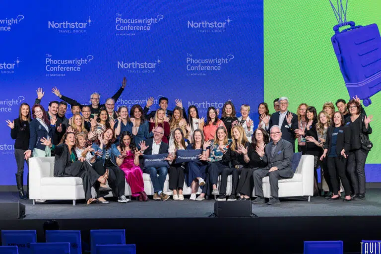 A large group of people, dressed in business and casual attire, smiling and waving at the camera. They are gathered on stage with a blue backdrop featuring logos of Northstar Travel Group and the Phocuswright Conference.