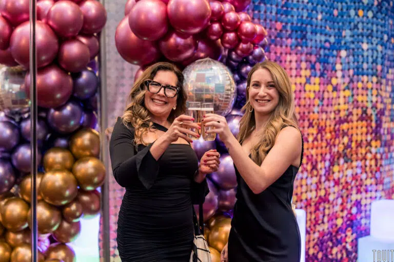 Two women smiling and raising glasses in front of colorful balloon decorations and a pixelated backdrop. They are dressed in elegant attire.