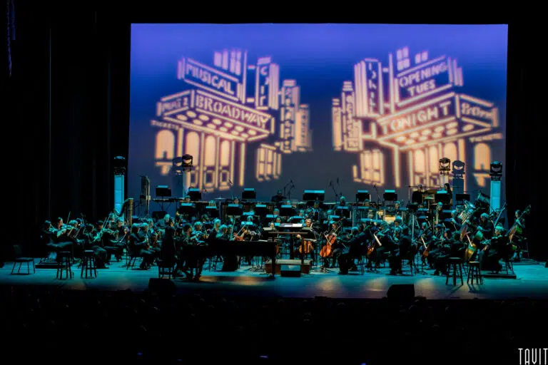 An orchestra performs on stage with a backdrop depicting stylized Broadway signs. The stage is lit in blue, and various musicians are seated with their instruments, creating a dramatic and theatrical atmosphere.