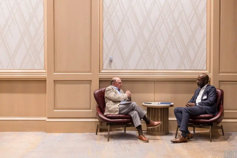 Two men sitting on leather chairs in a spacious room with patterned walls. A small table with a notebook and pen separates them. Both are engaged in conversation, one gesturing with his hands while the other listens attentively.