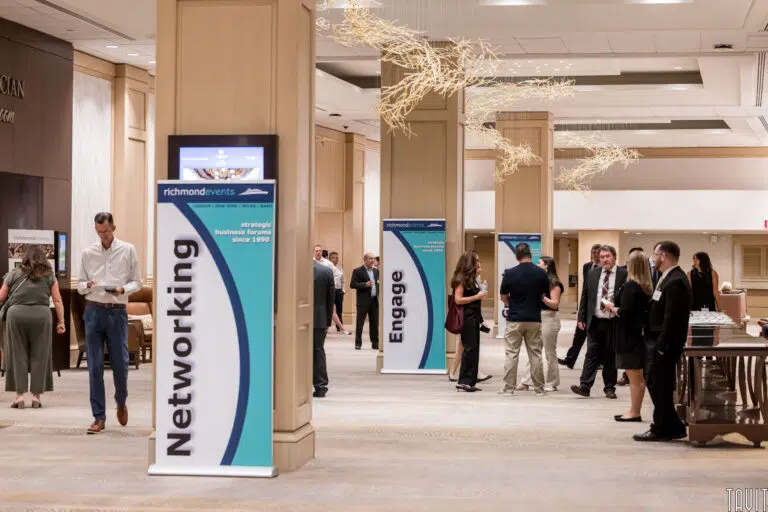 People networking in a conference hall with large pillars and banners reading "Networking" and "Engage." The setting is bright and modern, with a few people conversing in small groups.