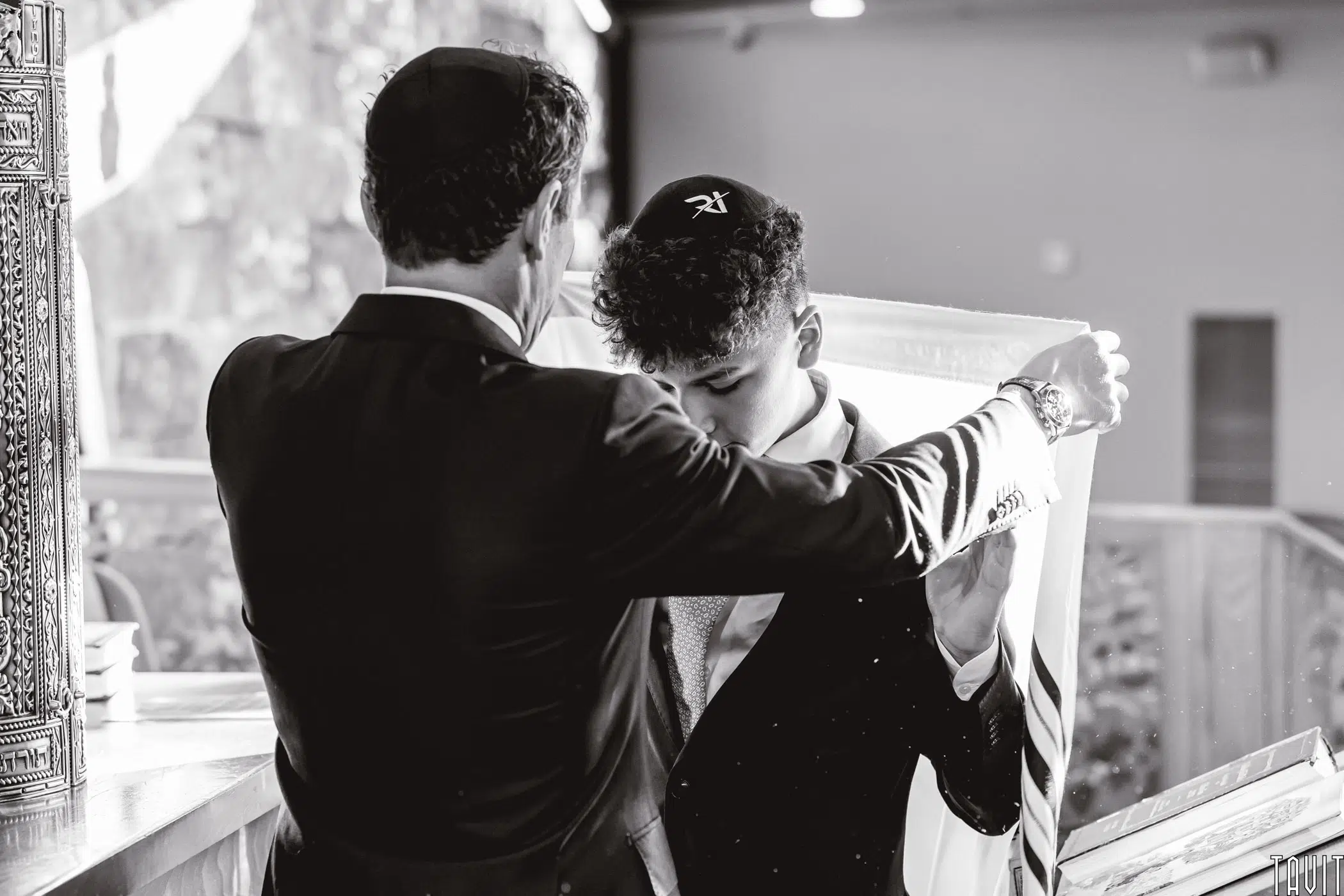 A black-and-white image of a man draping a tallit over a young man's shoulders in a synagogue setting. Both are wearing kippahs. The scene conveys a sense of tradition and ceremony.