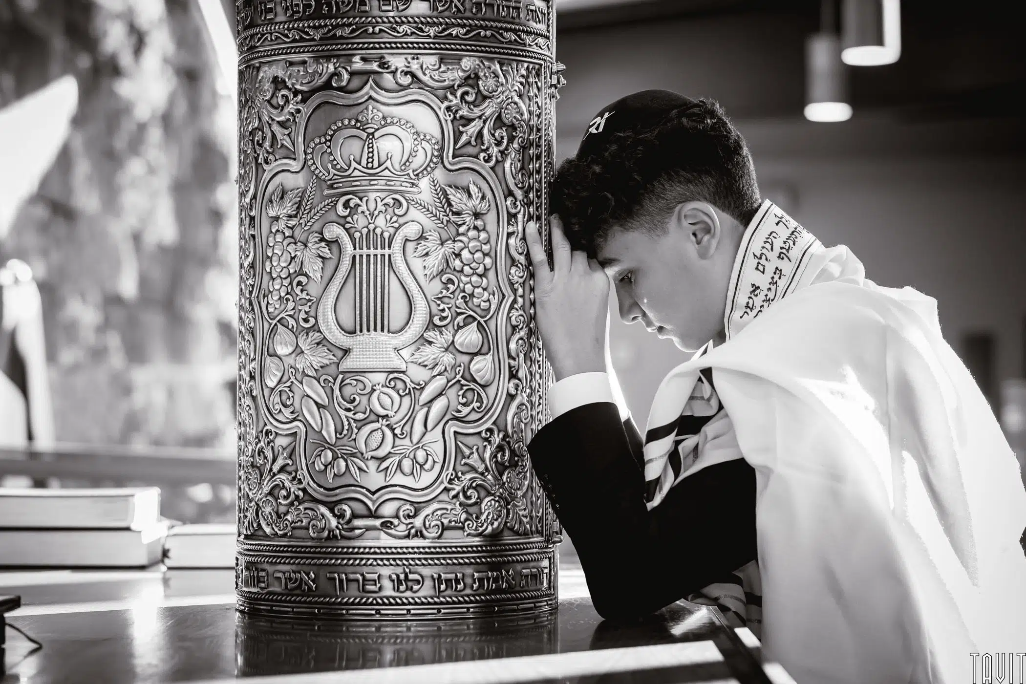 A young person in prayer, wearing a tallit, leans their head against a large, ornate Torah ark adorned with intricate designs. The setting is a well-lit room, and the atmosphere is reflective and peaceful.