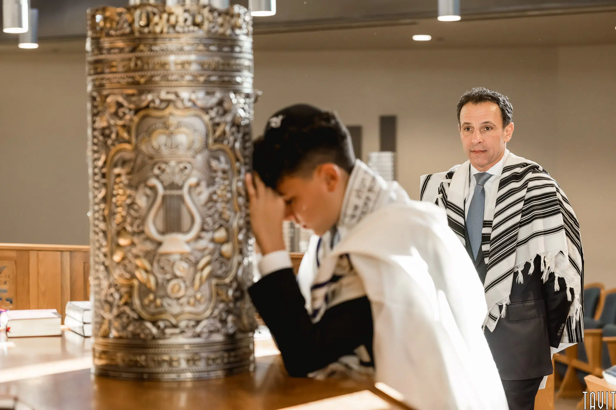A young boy wearing a tallit stands in front of a Torah ark, with his head bowed and hands covering his face. An older man, also wearing a tallit, stands behind him. The setting appears to be a synagogue.