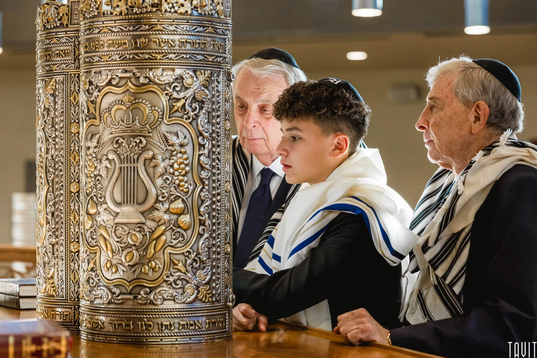Three men of different ages stand closely together, wearing traditional Jewish prayer shawls and kippahs, near an ornate Torah case. They appear to be engaged in a religious ceremony inside a synagogue.