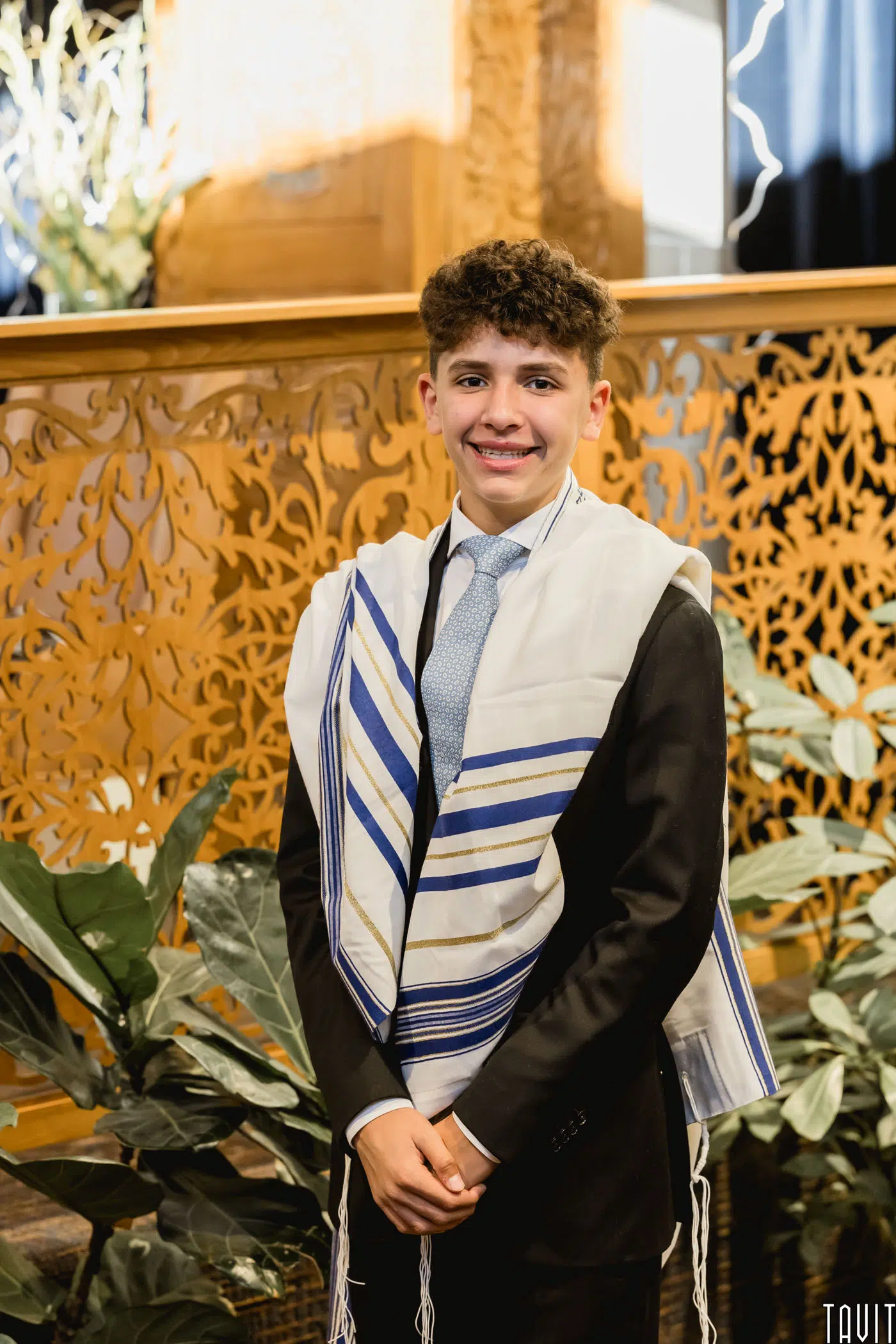 A young person wearing a suit, blue tie, and a white prayer shawl with blue stripes stands smiling in front of an ornate wooden structure with leafy green plants around.