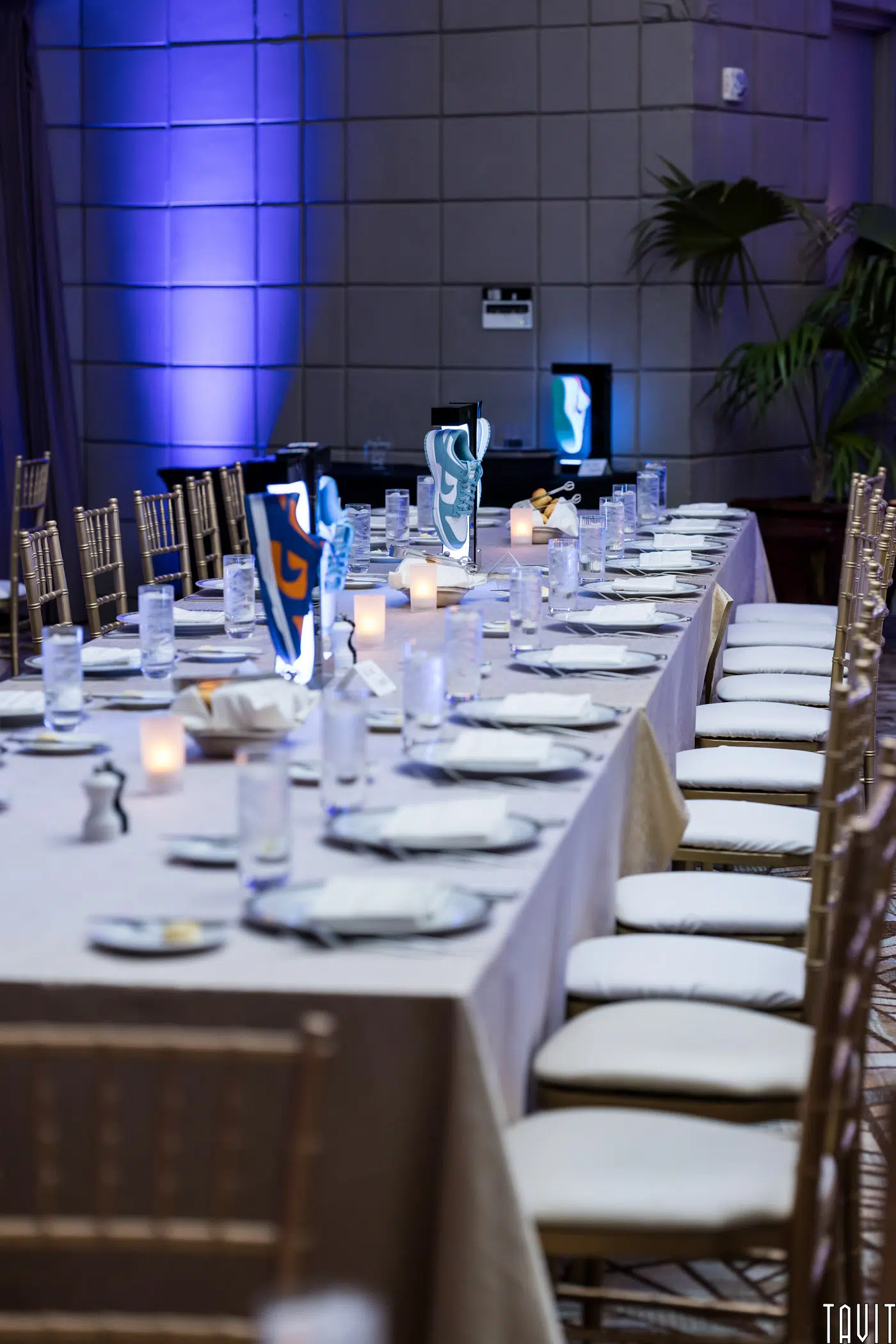 A long dining table set for an event, covered with a beige tablecloth. It features rows of white plates, glasses, and folded napkins. Candles in glass holders are lit, and two illuminated decorative panels with abstract designs are visible in the background.