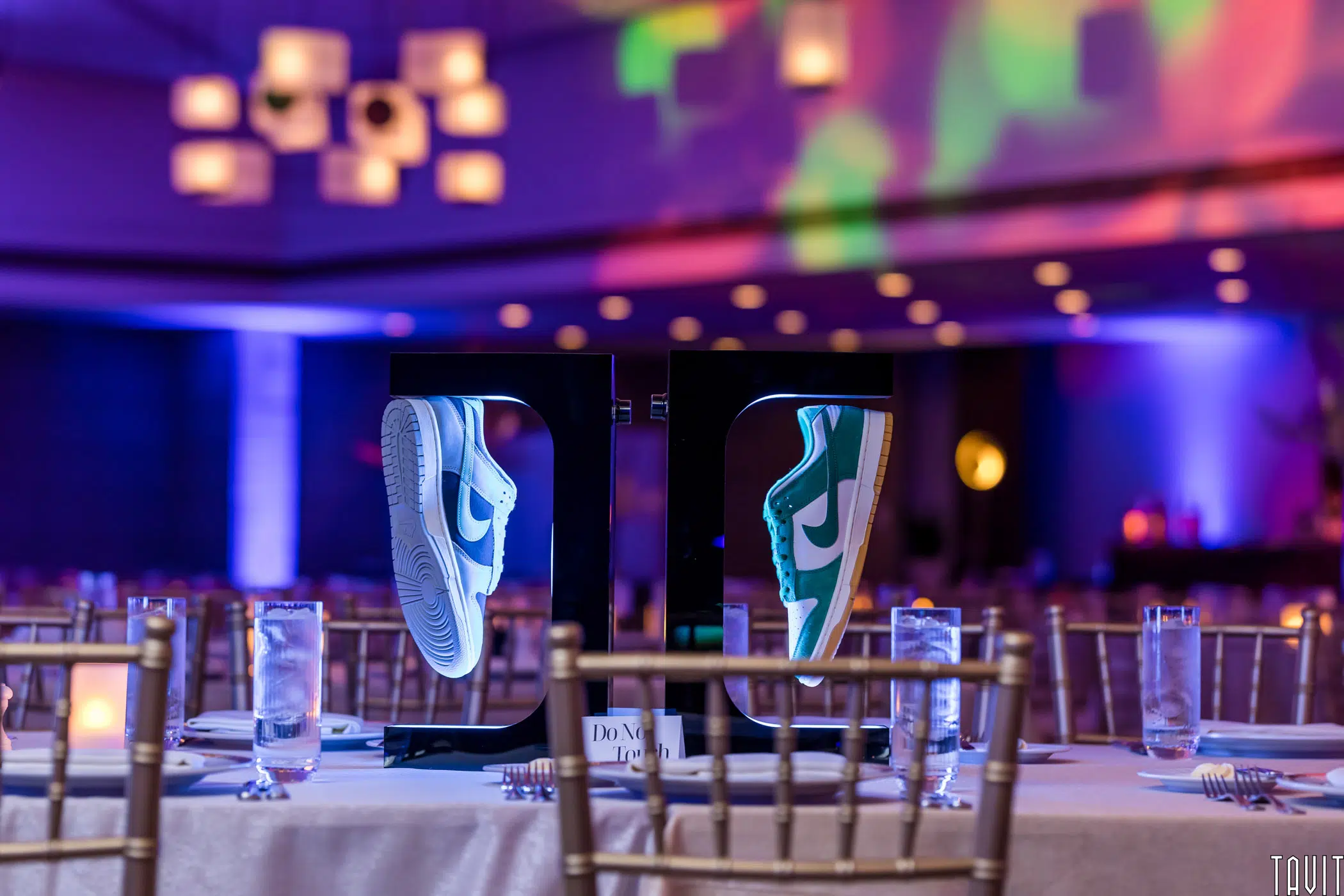 A pair of sneakers displayed uniquely, appearing to float in frames, on a table set for an event. The background features colorful lighting and elegant chandeliers, creating a vibrant and sophisticated atmosphere.