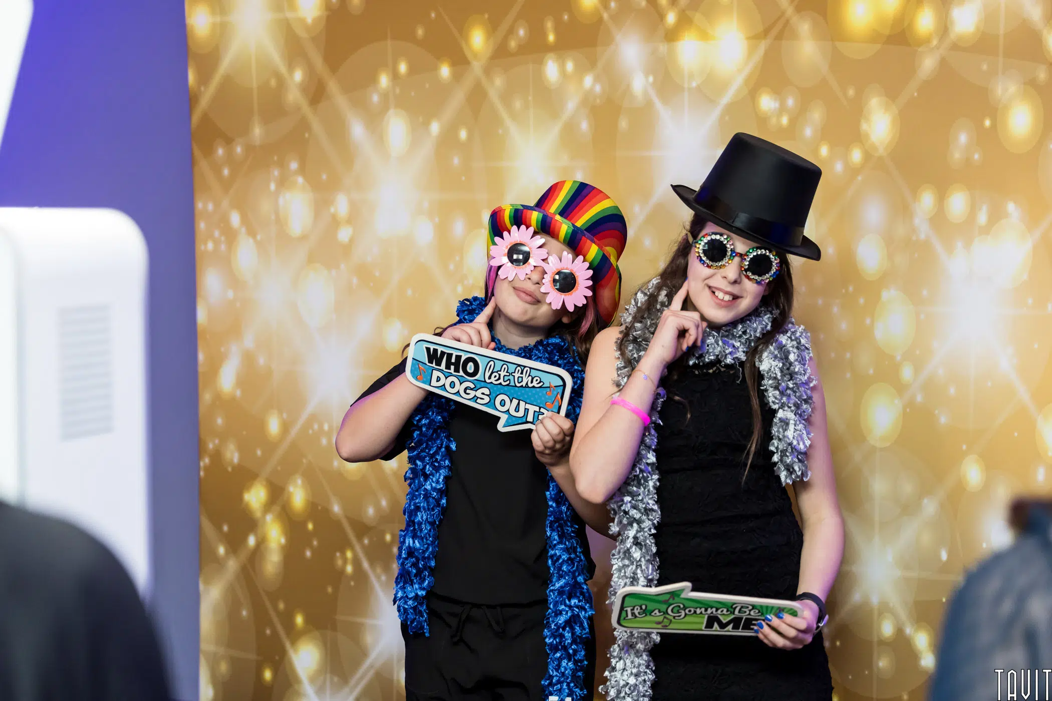 Two kids dressed in colorful hats and sunglasses pose in front of a festive backdrop. One holds a sign that says "WHO let the DOGS OUT," and the other holds a sign that says "Let’s Groove Tonight." They both wear shiny scarves.