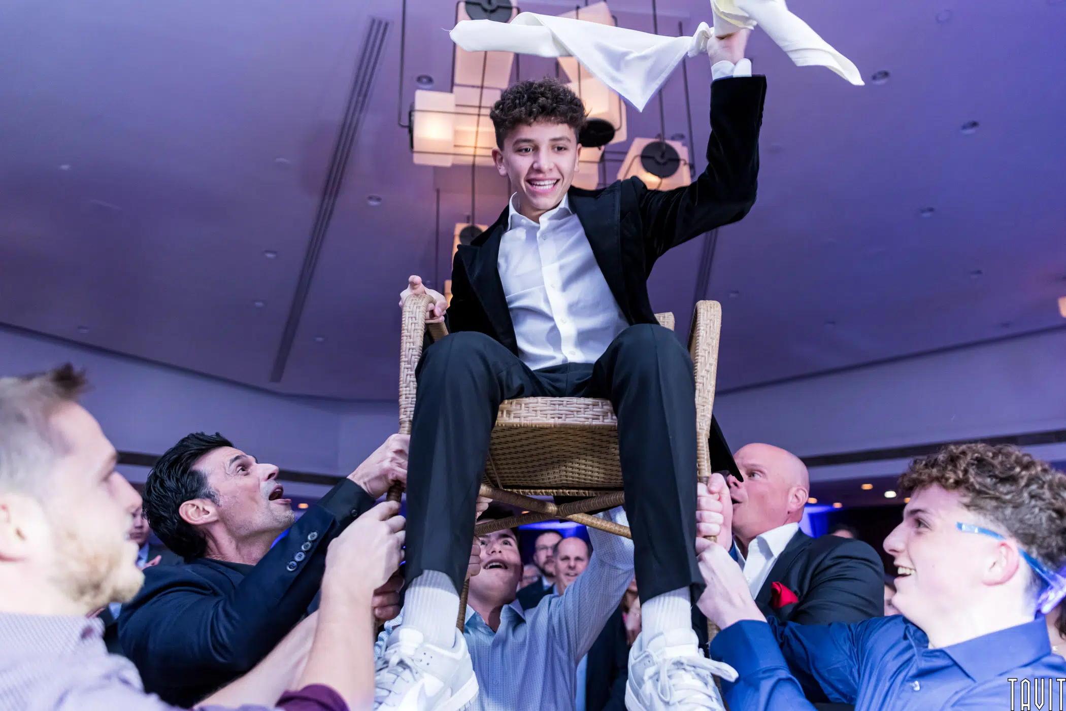 A young man in a suit is joyfully lifted on a chair by several people during a celebration. He holds a napkin in one hand, grinning, as the energetic crowd supports him. The setting appears festive, with a bright, cheerful atmosphere.