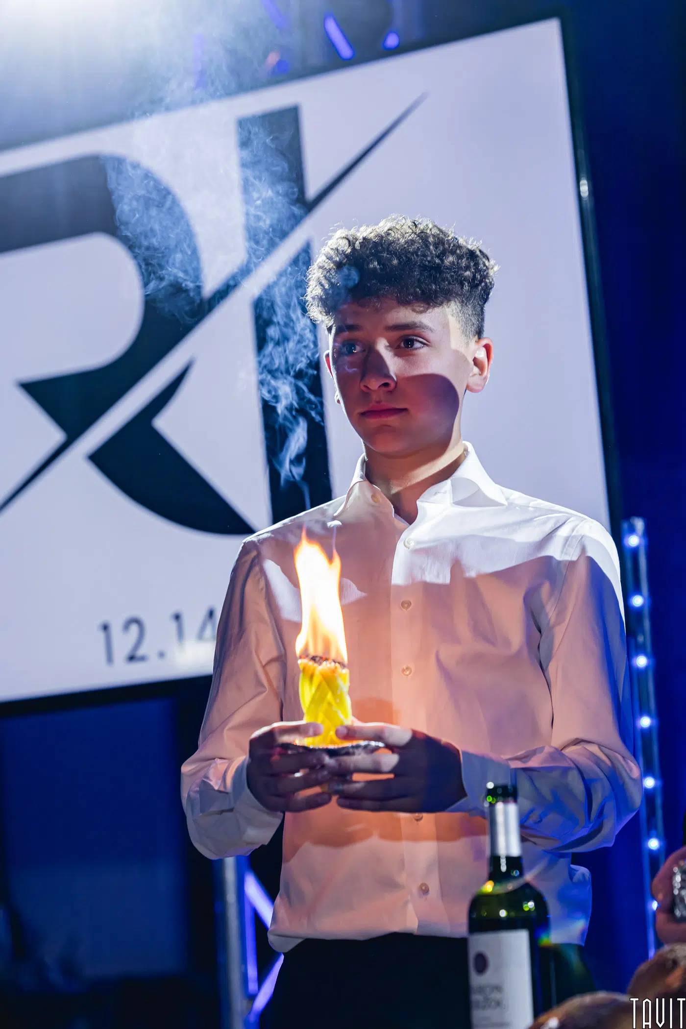 A young man in a white shirt holds a flaming dessert on a plate. He stands in front of an illuminated sign with abstract letters and a date. A wine bottle is on the table in front of him. The setting appears to be a formal event.