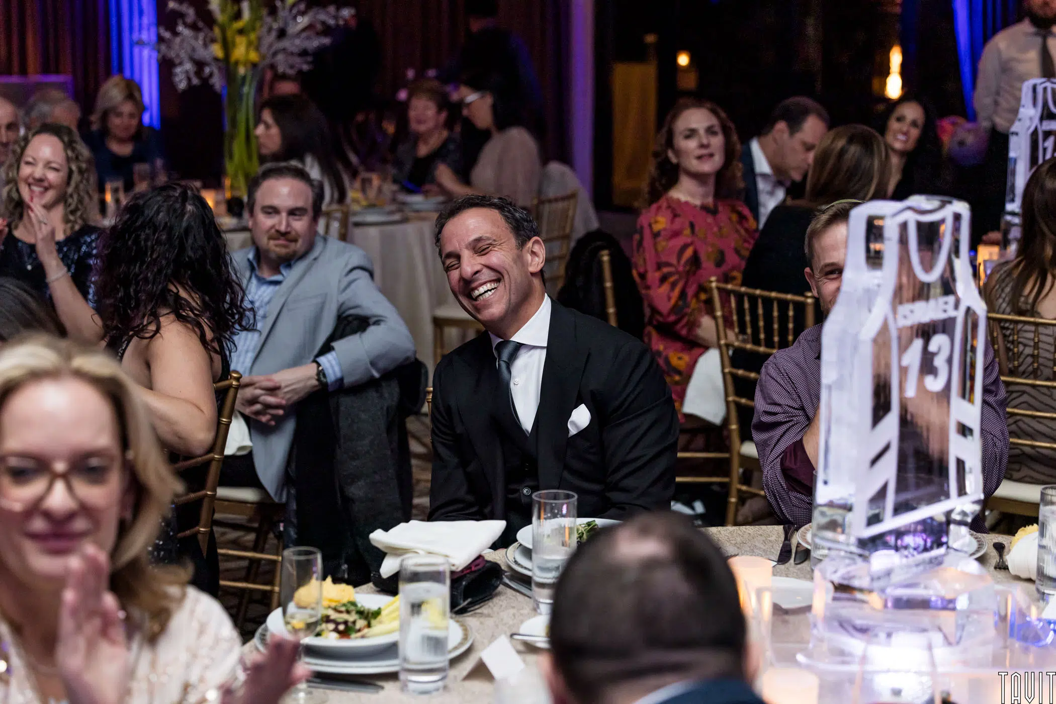 A joyful man in a suit sits at a round table, surrounded by people dining and talking in a warmly lit event setting. In the foreground, an ice sculpture of a sports jersey with the number 13 is prominent.